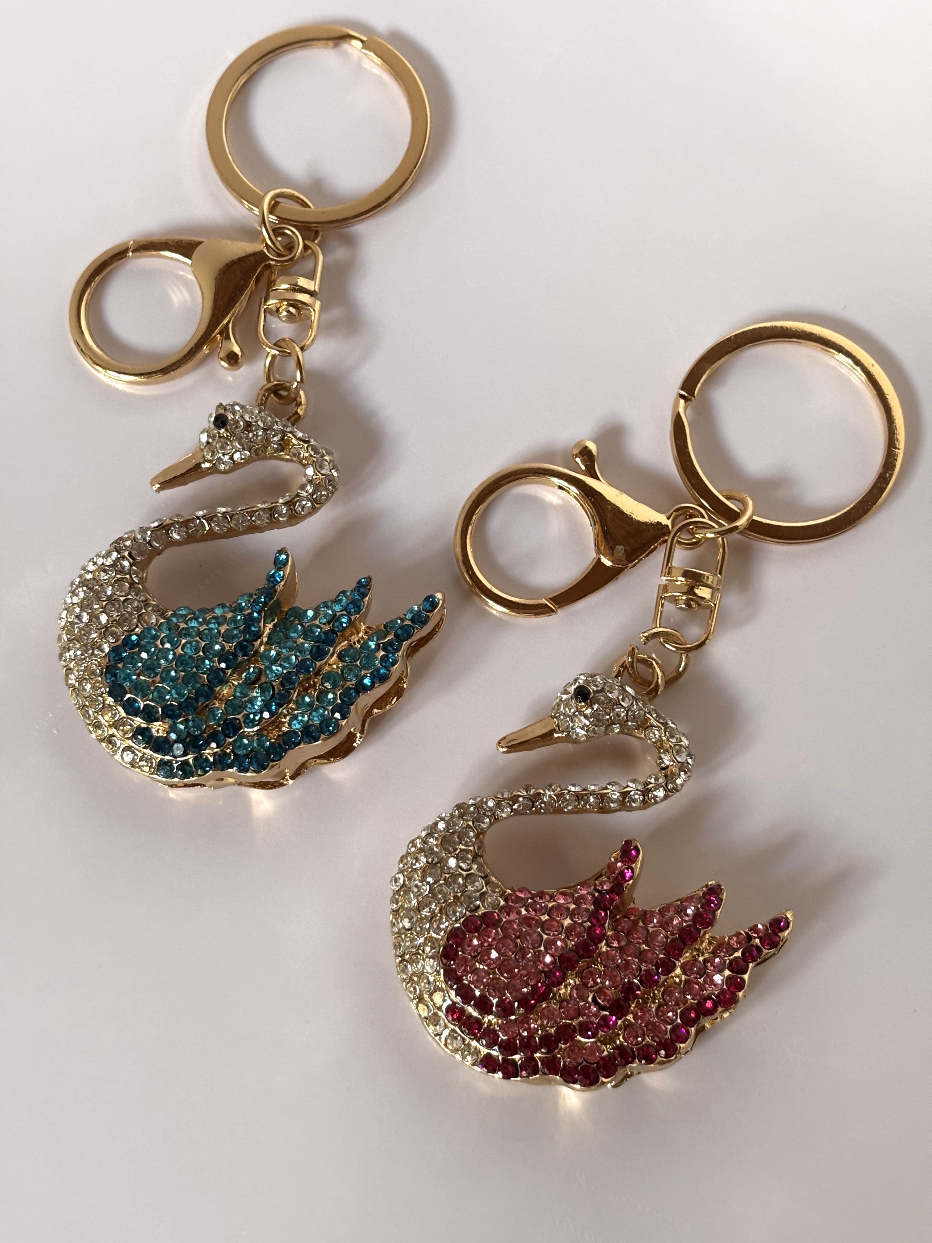 Due portachiavi gioiello a forma di cigno con pavé di strass brillanti e ali sfumate in cristalli colorati, uno nei toni dell'azzurro oceano e uno nei toni del rosa magenta, con ganci dorati su sfondo bianco.