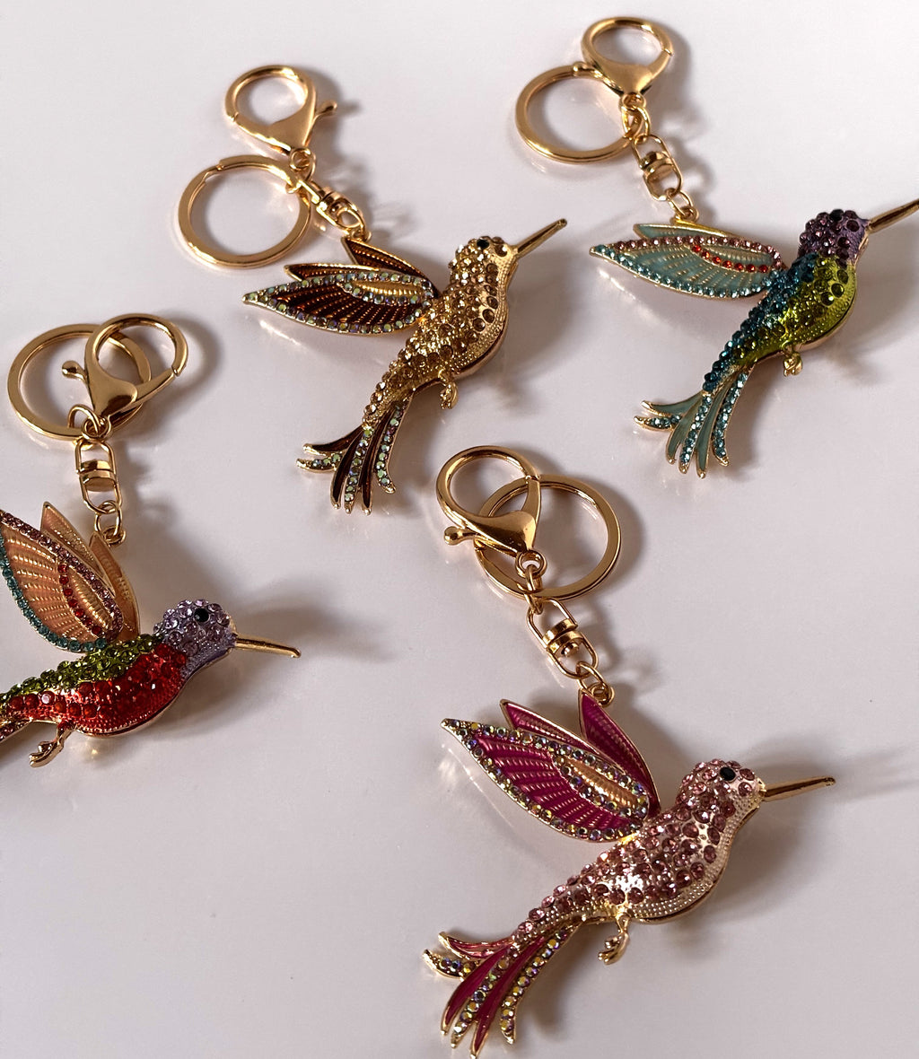 Quattro portachiavi gioiello a forma di colibrì tempestati di strass lucenti in varianti cromatiche assortite tra cui oro, viola-verde, arancio-verde e rosa-magenta, con agganci dorati.