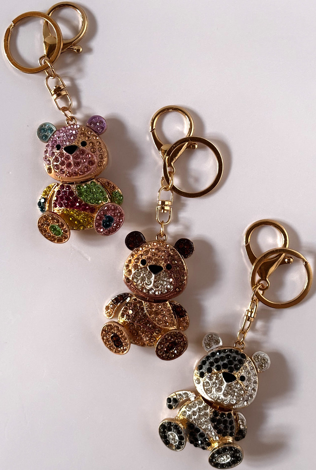 Tre portachiavi gioiello a forma di orsetto tempestati di strass lucenti con design patchwork geometrico, disponibili nelle varianti multicolore arcobaleno, bronzo ambrato e bianco-nero panda, con ganci dorati.