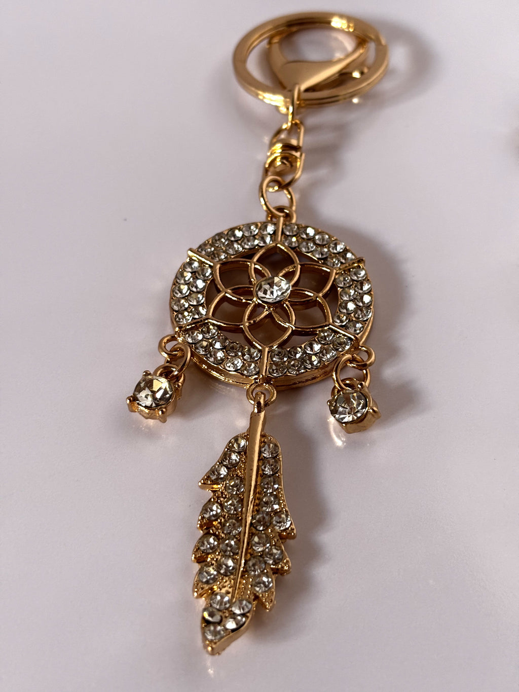 Portachiavi gioiello a forma di acchiappasogni con piuma pendente tempestato di strass lucenti, disponibile nelle varianti di colore bianco trasparente, azzurro turchese e rosa magenta su struttura in lega metallica dorata.