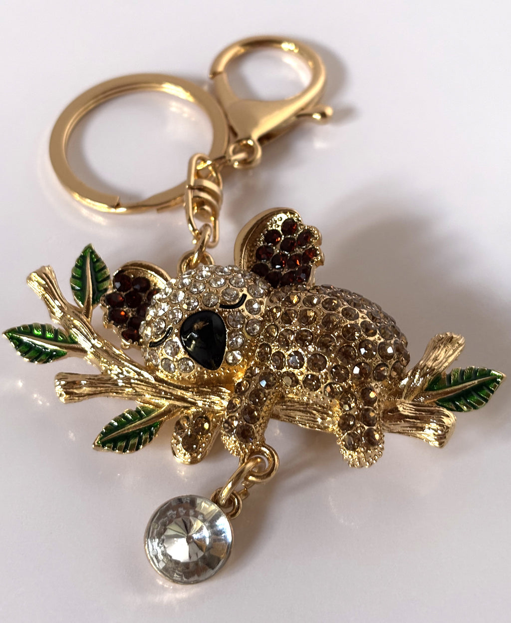 Portachiavi gioiello tridimensionale a forma di koala tempestato di strass lucenti color ambra e bianco, aggrappato a un ramo dorato con foglie smaltate verdi e un grande cristallo pendente.