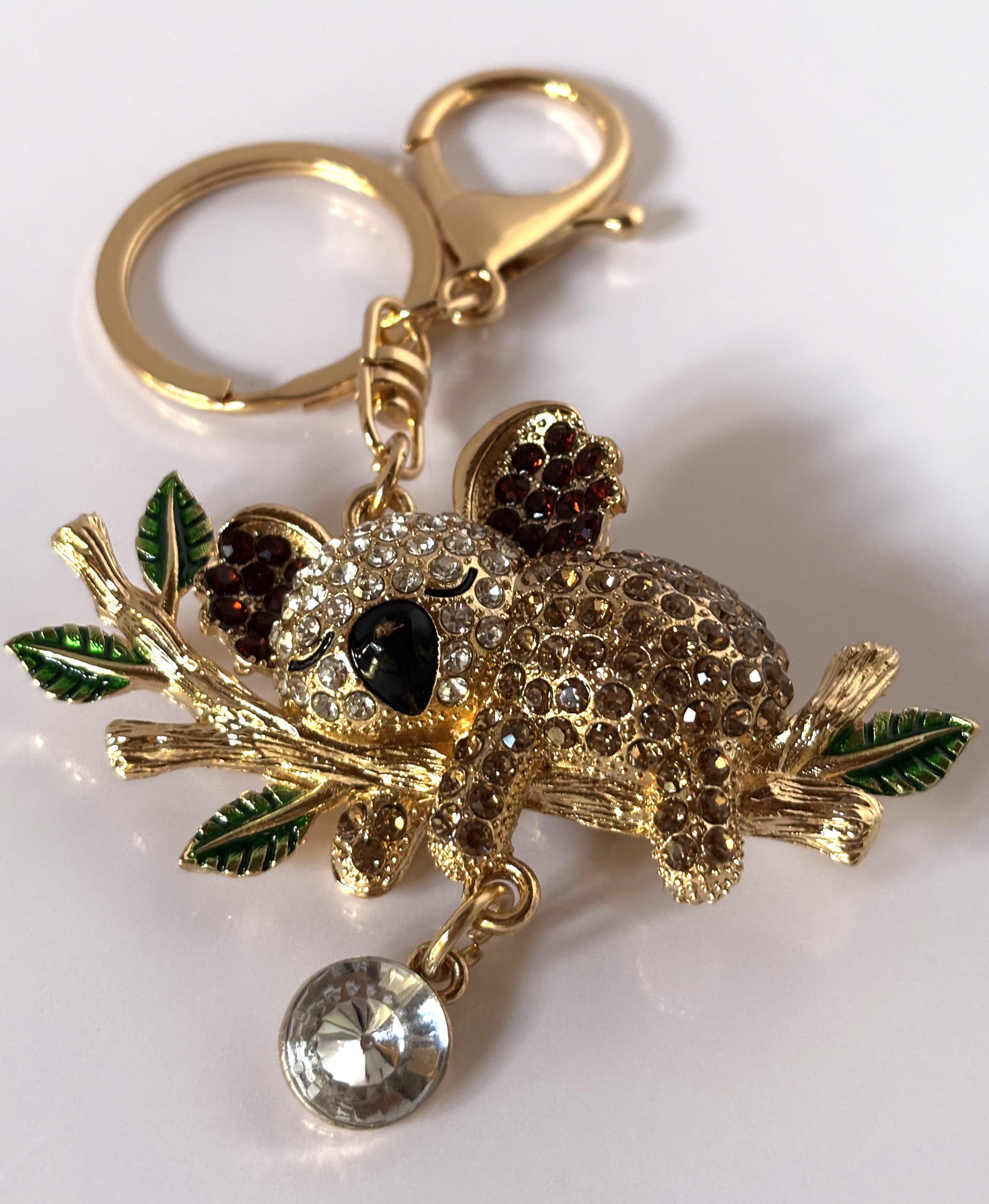 Portachiavi gioiello tridimensionale a forma di koala tempestato di strass lucenti color ambra e bianco, aggrappato a un ramo dorato con foglie smaltate verdi e un grande cristallo pendente.