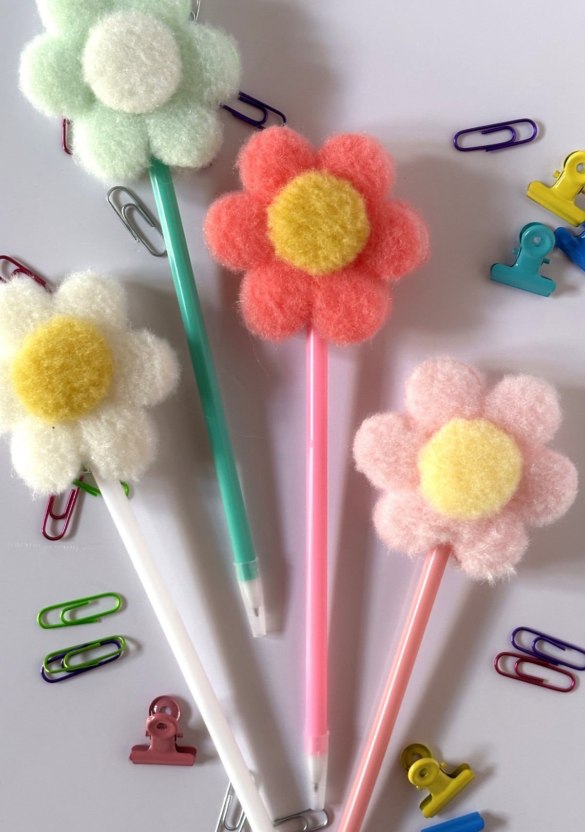 Set completo di quattro penne con topper tridimensionale a forma di margherita in tessuto morbido pon pon nei colori verde menta, fucsia, bianco e rosa pastello, circondate da graffette e clip colorate.