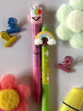 Set composto da due penne in stile Kawaii, una rosa con topper a tazza unicorno e una verde con topper tridimensionale ad arcobaleno, disposte accanto ad accessori per ufficio colorati.