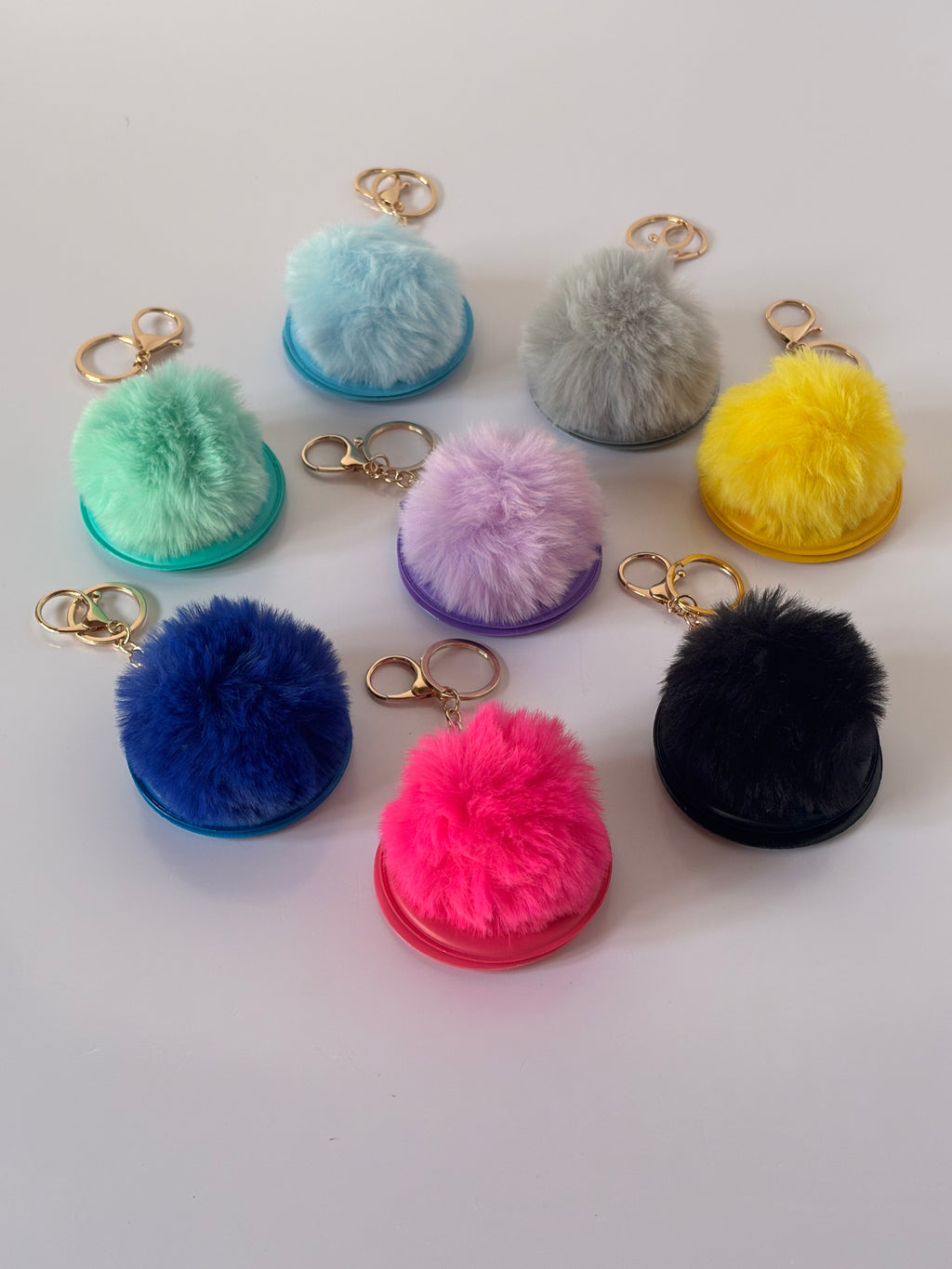 Vista dall'alto della collezione completa di portachiavi specchietto C.Moon disposti a cerchio nelle tonalità azzurro cielo, grigio perla, giallo sole, lilla, verde menta, blu elettrico, fucsia shocking e nero, evidenziando i soffici pompon e i moschettoni dorati.