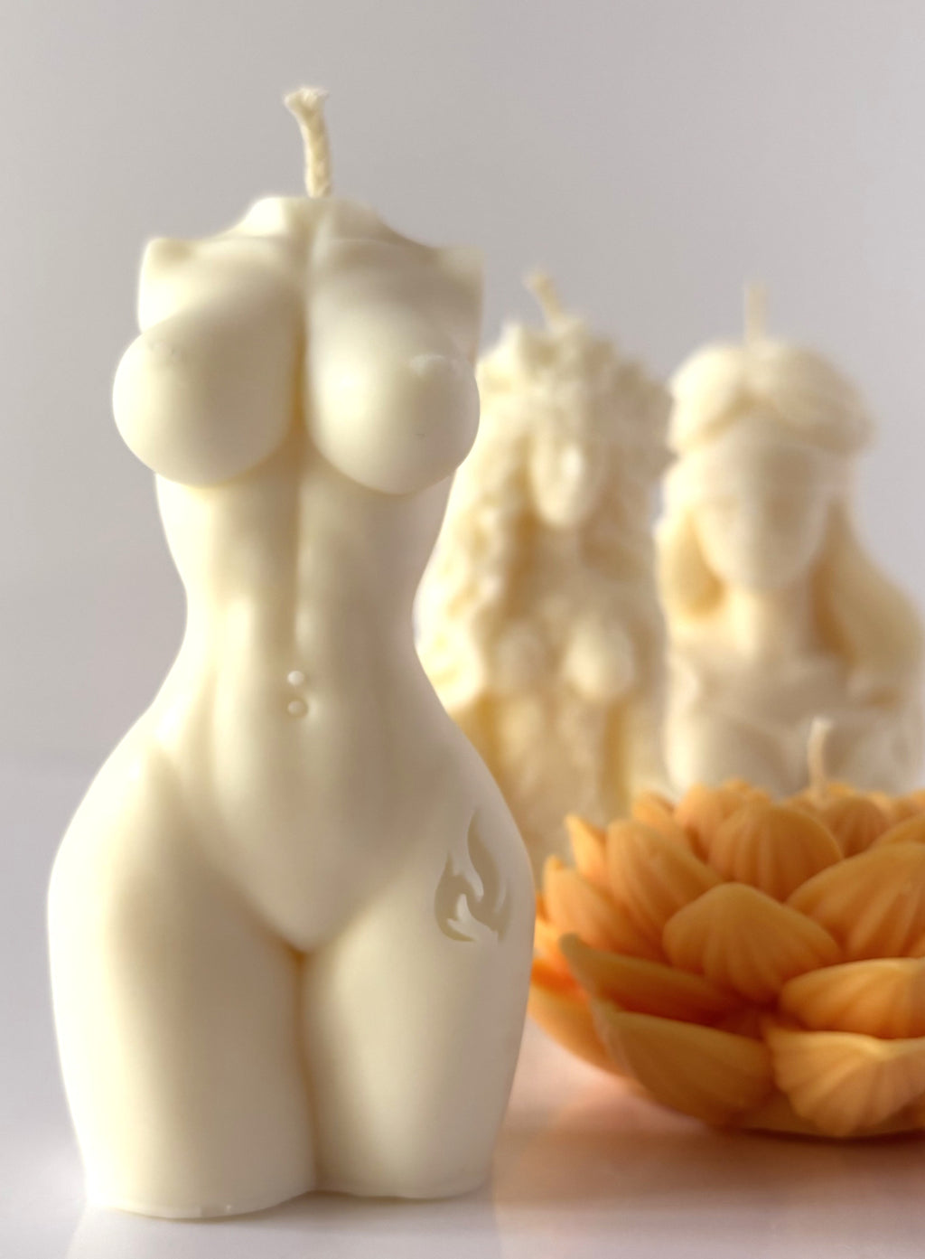 Candela scultorea C.Moon color bianco avorio a forma di busto e torso femminile con incisione a fiamma sulla coscia, con candele Dea Gaia e fiore di loto sfocate sullo sfondo.