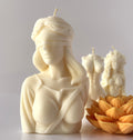 Candela scultorea C.Moon color bianco avorio raffigurante il busto della Dea della Giustizia bendata, con candele a torso e fiore di loto sfocate sullo sfondo.
