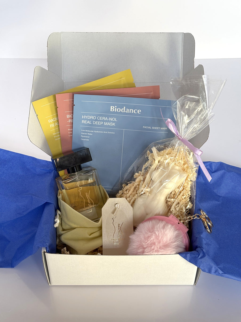 Cofanetto regalo Femme Box C.Moon da 59.90€ aperto su fondo bianco, contenente il profumo da 50ml Defining Your Glow, un tris di maschere Biodance (azzurra, rosa, gialla), la candela scultorea a forma di torso femminile Empower Body e il portachiavi specchietto rosa con pompon.