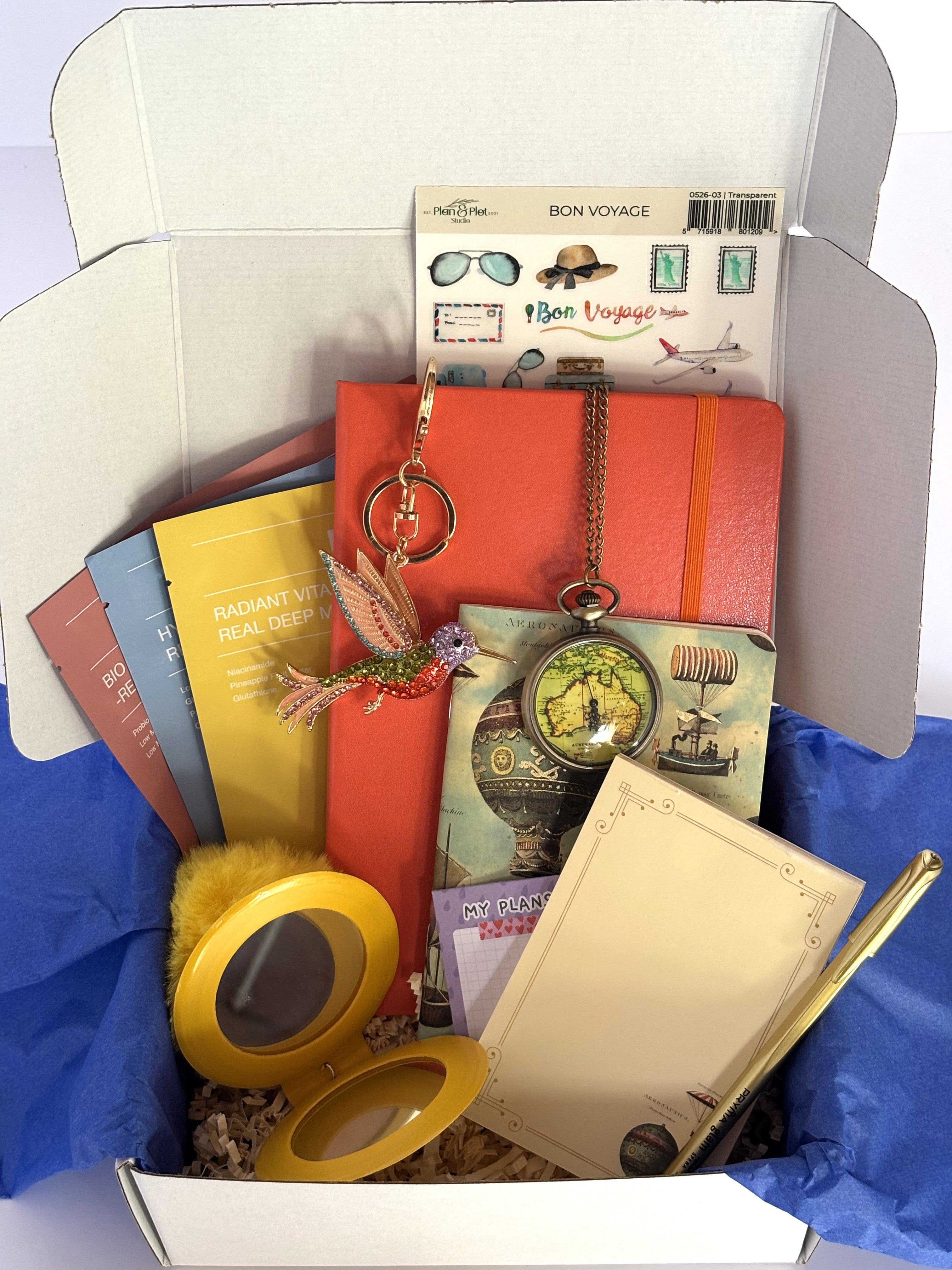 Cofanetto regalo Travel Box C.Moon da 69.90€ aperto su fondo bianco con velina blu, contenente un taccuino rigido arancione, un taccuino vintage Kartos con mongolfiere, un orologio da taschino antico, un portachiavi a forma di colibrì glitterato, quattro maschere Biodance, una penna stilografica dorata e un portachiavi specchietto giallo sole