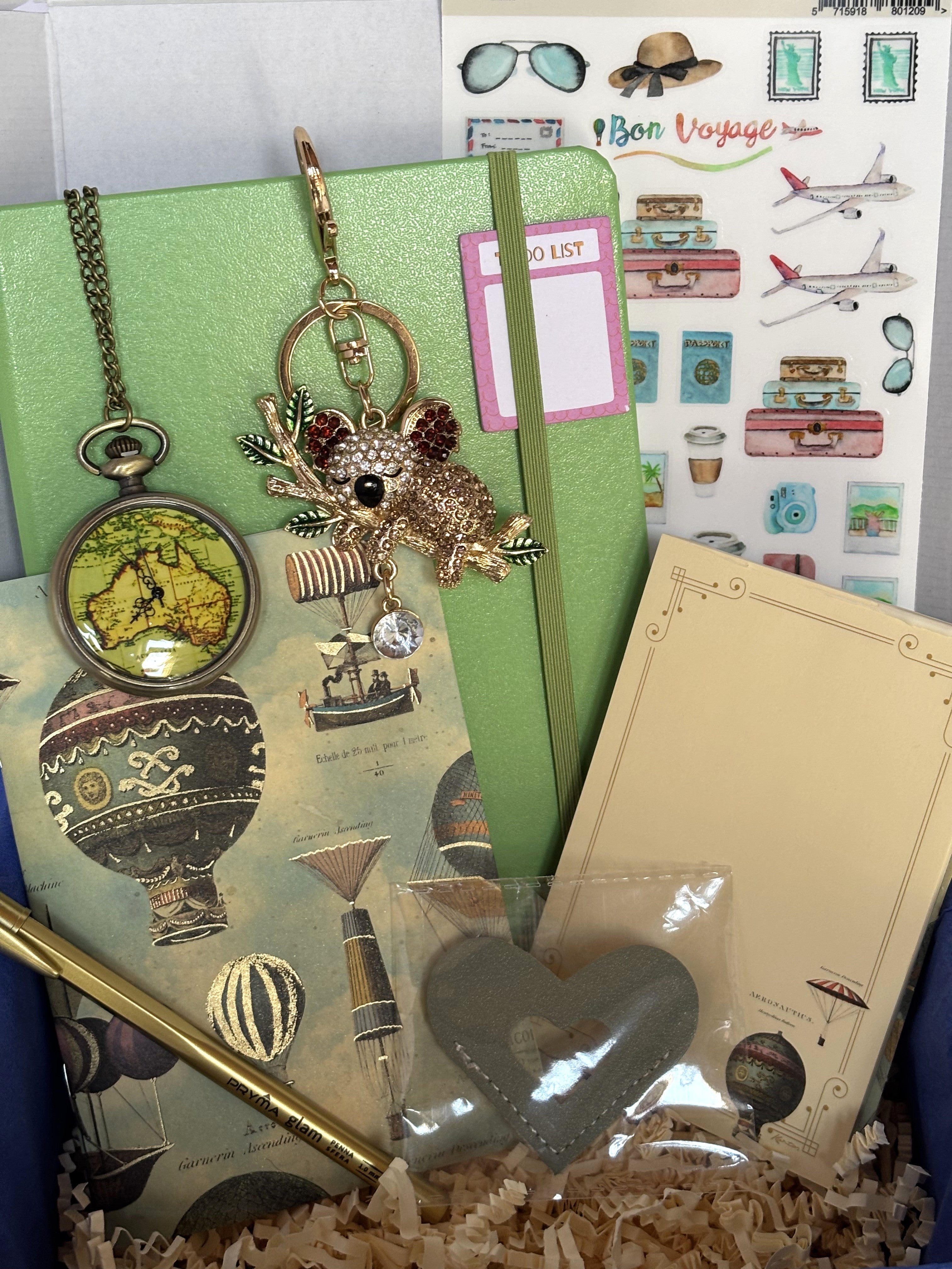 Cofanetto regalo Travel Box C.Moon da 49.90€ variante verde aperto su fondo bianco con velina blu, contenente un taccuino rigido verde mela, taccuino Kartos con mongolfiere, orologio da taschino, portachiavi a koala con strass, penna dorata e segnalibro a cuore grigio