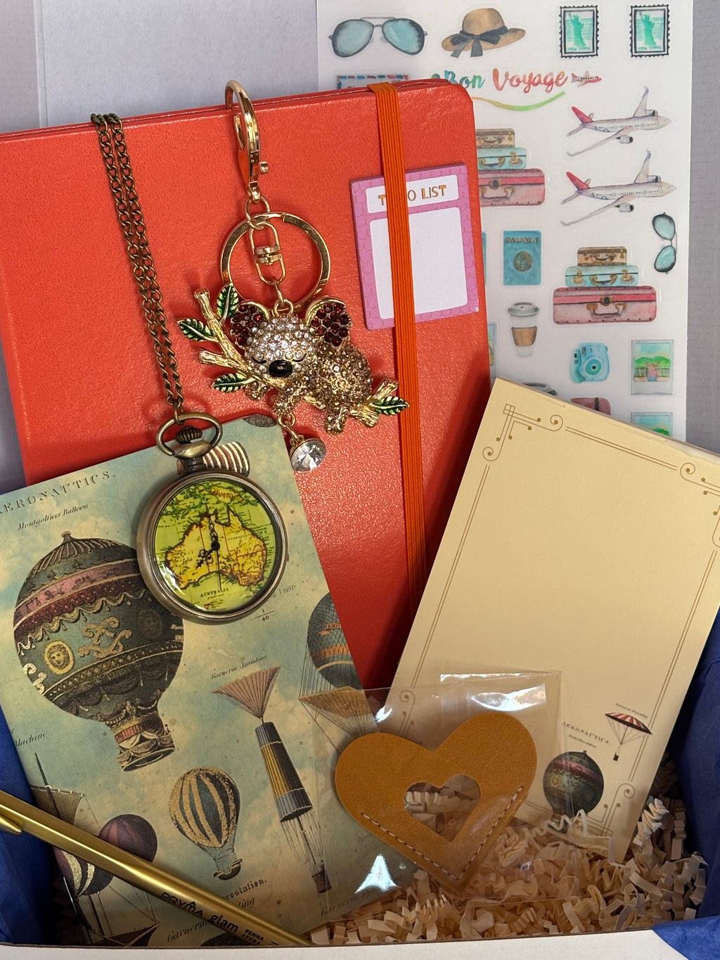 Cofanetto regalo Travel Box C.Moon da 49.90€ variante arancione aperto su fondo bianco con velina blu, contenente un taccuino rigido arancione, taccuino Kartos con mongolfiere, orologio da taschino, portachiavi a koala con strass e penna dorata