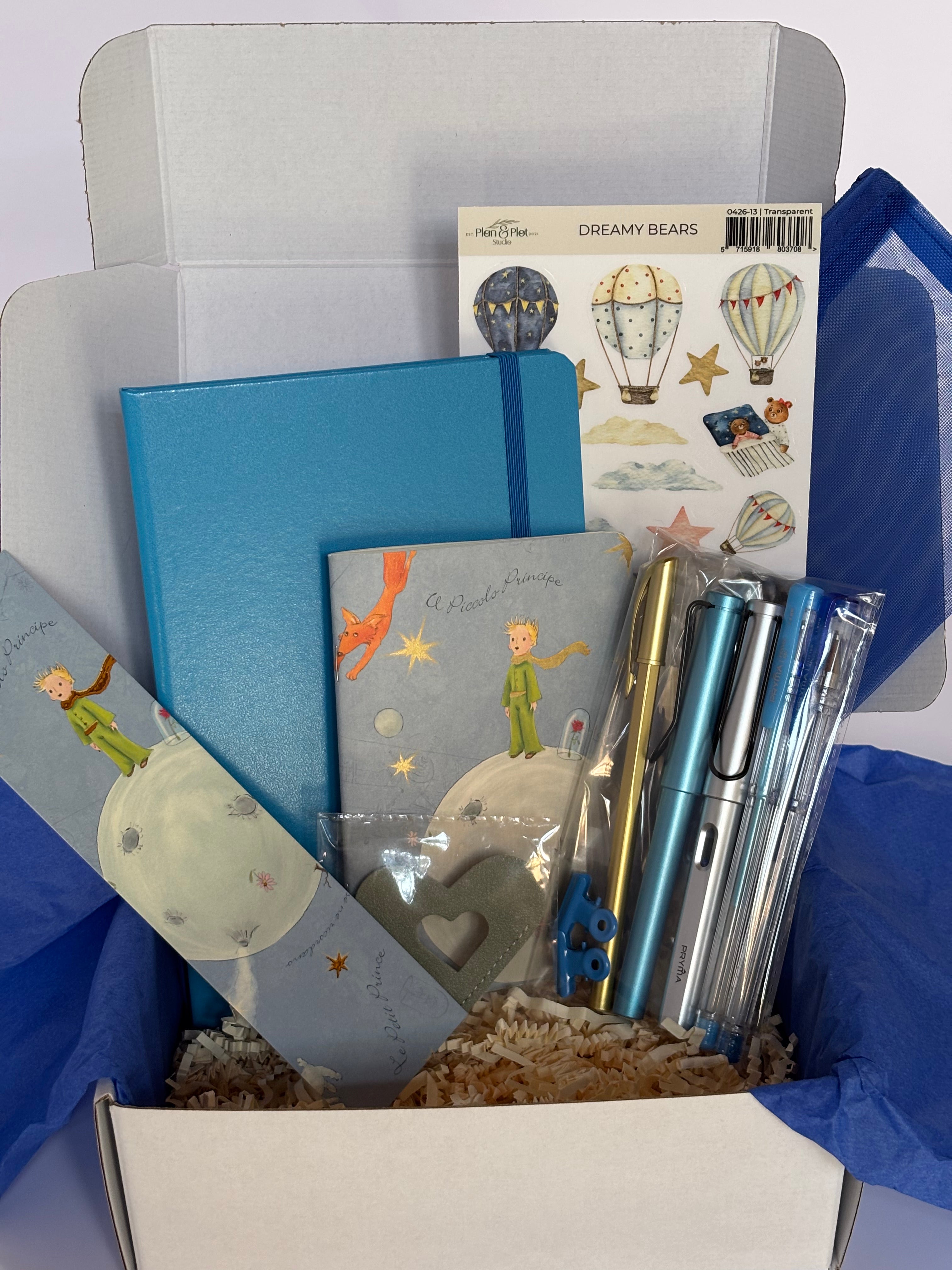 Cofanetto regalo Il Piccolo Principe C.Moon da 39.90€ aperto su fondo bianco con velina blu, contenente un taccuino rigido azzurro avio, un quaderno e un segnalibro del Piccolo Principe, una penna stilografica dorata, tre penne Pryma Trio, un astuccio blu in mesh e stickers con mongolfiere.